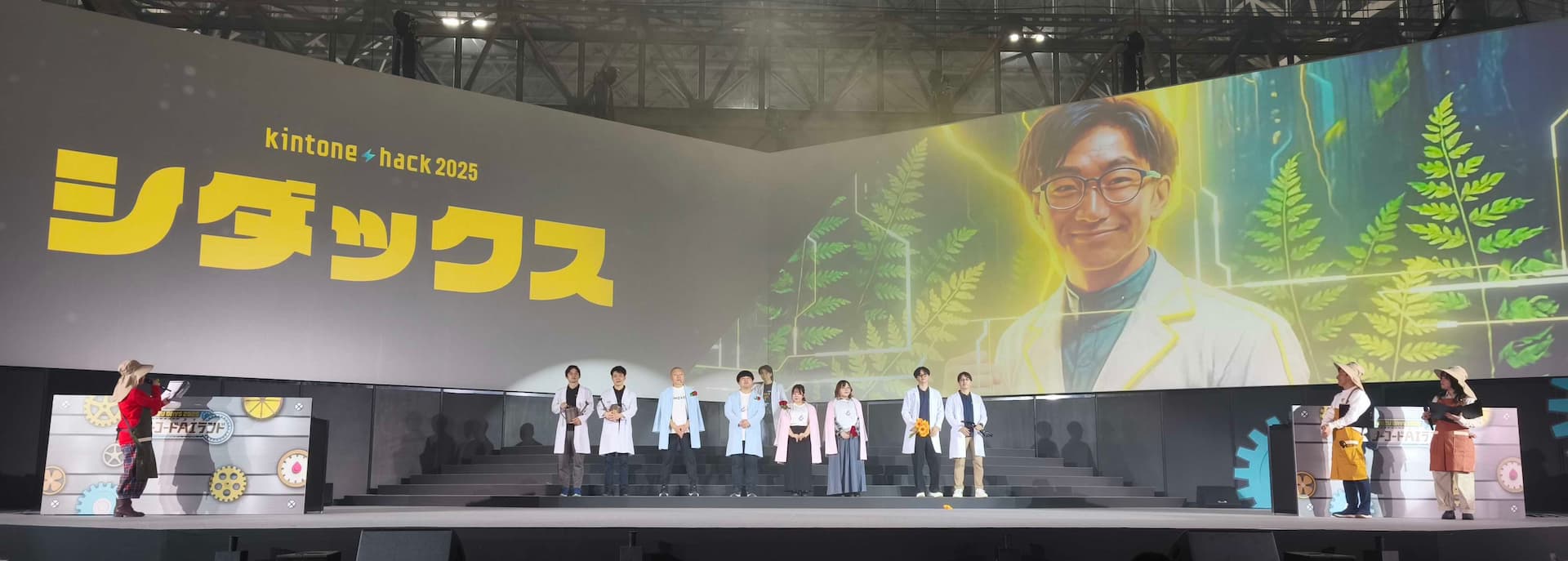 本戦の会場に登場したときの写真。巨大なスクリーンに「シダックス」と表示され、登壇者である志太悠真の顔写真をAIでイラスト化したものが映っている。ステージには登壇者たちが並んでいる。